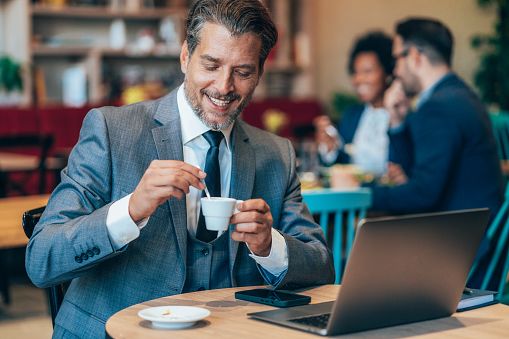 Lächelnder Mann um die vierzig in einem Café mit Laptop und Kaffeetasse, der eine ernsthafte Begegnung, einen positiven ersten Eindruck und authentische Kommunikation veranschaulicht.